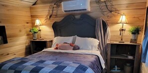 A wooden room with a bed, two bedside tables with lamps, and an air conditioning unit. - Cabin #1 Cozy "fishing" themed cabin by the creek. Recently featured in magazine (Linden)