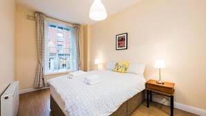 Une chambre à coucher avec un lit, des lampes de chevet, une fenêtre avec des rideaux, un radiateur et un tableau encadré au mur.