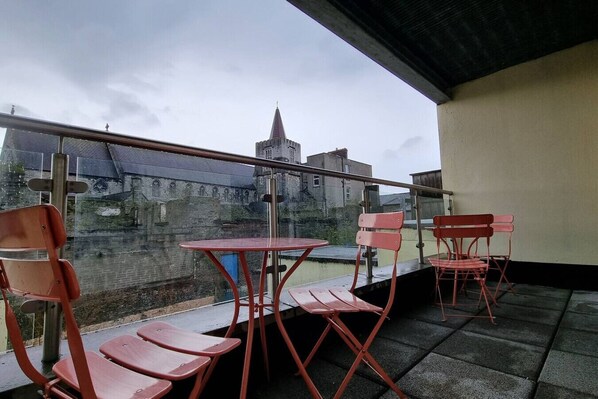 A balcony with red chairs and a table, overlooking a historic building with a tower.