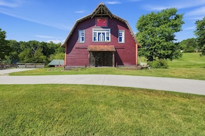 A red barn with a large door and windows, surrounded by a grassy field and trees.