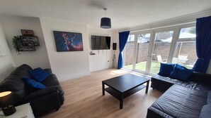 A living room with a black sofa, a coffee table, a TV, and a large window with curtains. - 3 BEDROOM + LIVING, Terrace House, Sleeps up to 7 people  with Garden (Cardiff)