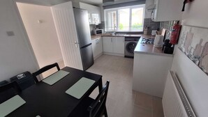 A compact kitchen with a refrigerator, microwave, sink, and a small dining area with a table and chairs. - 3 BEDROOM + LIVING, Terrace House, Sleeps up to 7 people  with Garden (Cardiff)