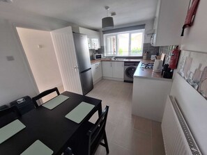 Een kleine keuken met een koelkast, magnetron, wastafel en een eethoekje met een tafel en stoelen.