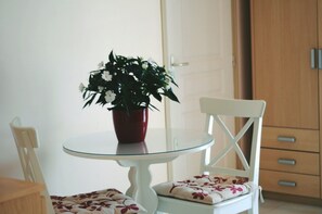 A dining area with a round glass table, a white chair with a floral cushion, and a potted plant with white flowers.