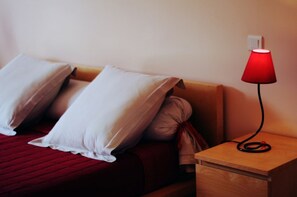 A bed with pillows, a red blanket, and a wooden nightstand with a red lamp.