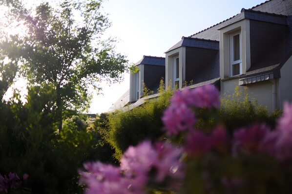 A residential building with a sloped roof, multiple windows, and surrounded by greenery.