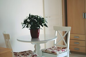 Une table à manger blanche avec un plateau en verre, une plante en pot rouge, une chaise blanche avec des coussins à motifs floraux, et une armoire en bois en arrière-plan.
