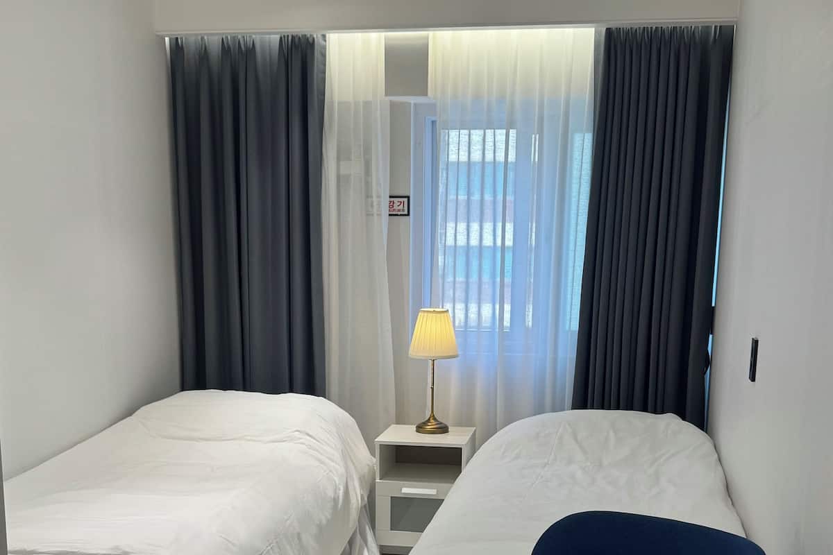 Une chambre d’hôtel avec deux lits, une petite table, une chaise, une lampe et une vue sur l’extérieur à travers des rideaux légers.