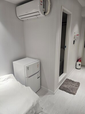 A small room with a bed, a white refrigerator, an air conditioner, a door, and a fire extinguisher.