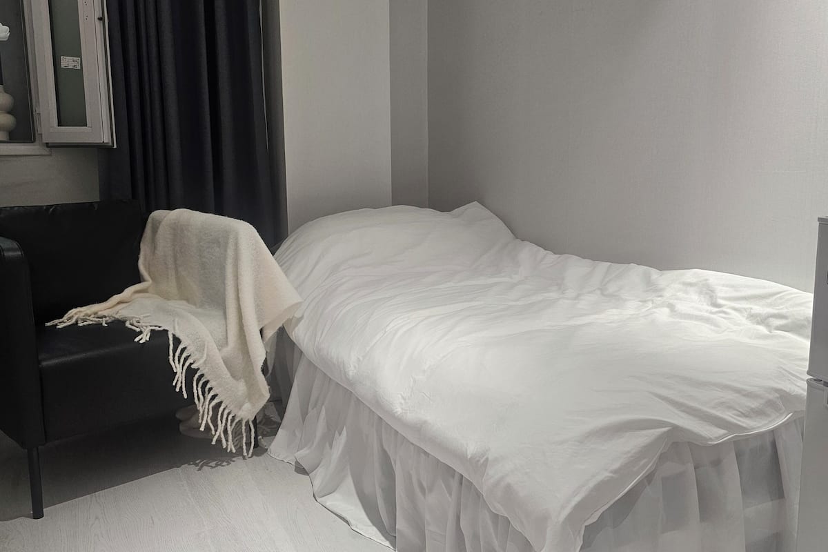 Une chambre avec un lit simple, une table de chevet, une chaise avec une couverture, une fenêtre avec des rideaux et un miroir.