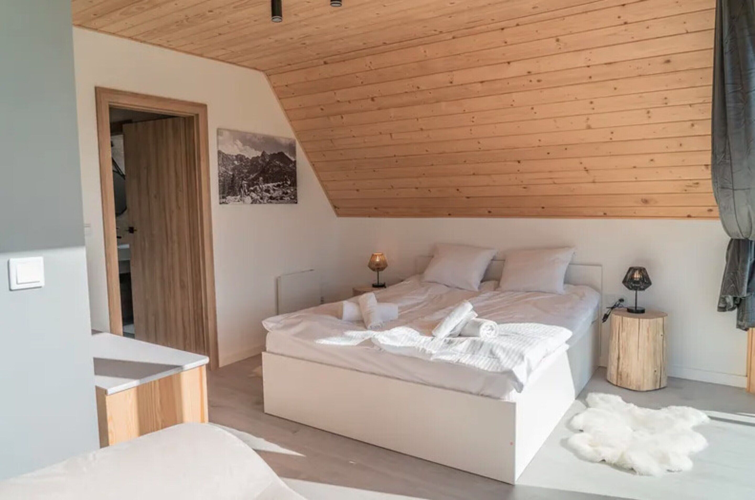 Un dormitorio con techo de madera, una cama con ropa blanca, una mesita de noche pequeña, una lámpara y una alfombra de piel.