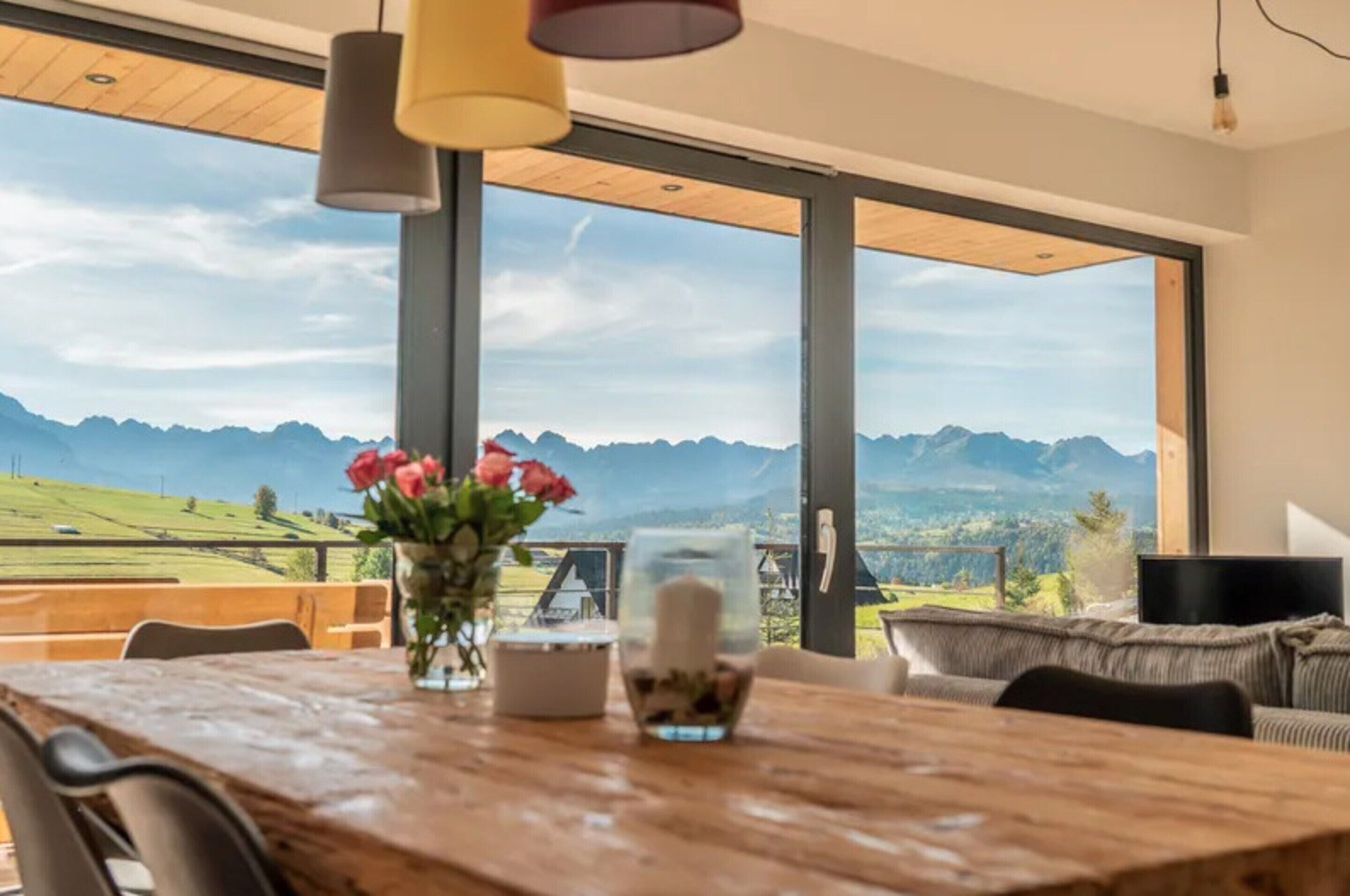 Uma sala de jantar com uma mesa de madeira, um vaso com flores e vista para montanhas através de grandes janelas.