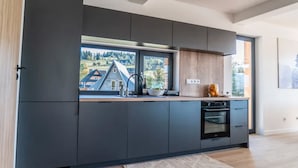 A modern kitchen with dark cabinetry, a large window with a view of a house, and a built-in oven. - Holiday Home With Sauna and Mountain View (Bukowina Tatrzanska)