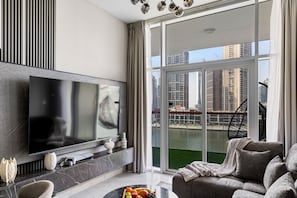Een moderne woonkamer met een grote flatscreen televisie, een bank, een salontafel met fruit en uitzicht op een stadsgezicht door grote ramen.
