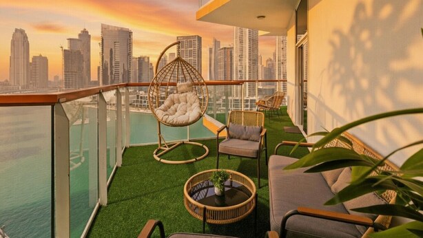 A balcony with a hanging chair, a sofa, and a small table, overlooking a cityscape at sunset. - Gogoto4 2BR Premium Canal & Burj Khalifa View (Dubai)