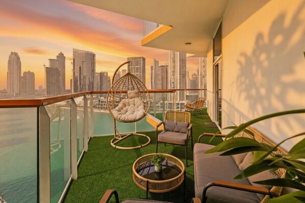A balcony with a hanging chair, a sofa, and a small table, overlooking a cityscape at sunset.