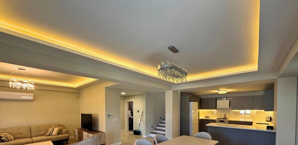 Una sala de estar moderna con un sofá, televisor, candelabro y una zona de cocina con encimera y armarios.
