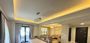 A modern hotel room with a large bed, a seating area with sofas, a chandelier, and a door.