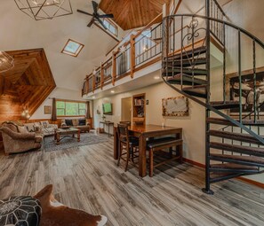 A spacious loft-style living area with a high ceiling, wooden floors, a central seating area with a sofa and chairs, a dining table, and a spiral staircase. - Gateway to Glacier (Columbia Falls)