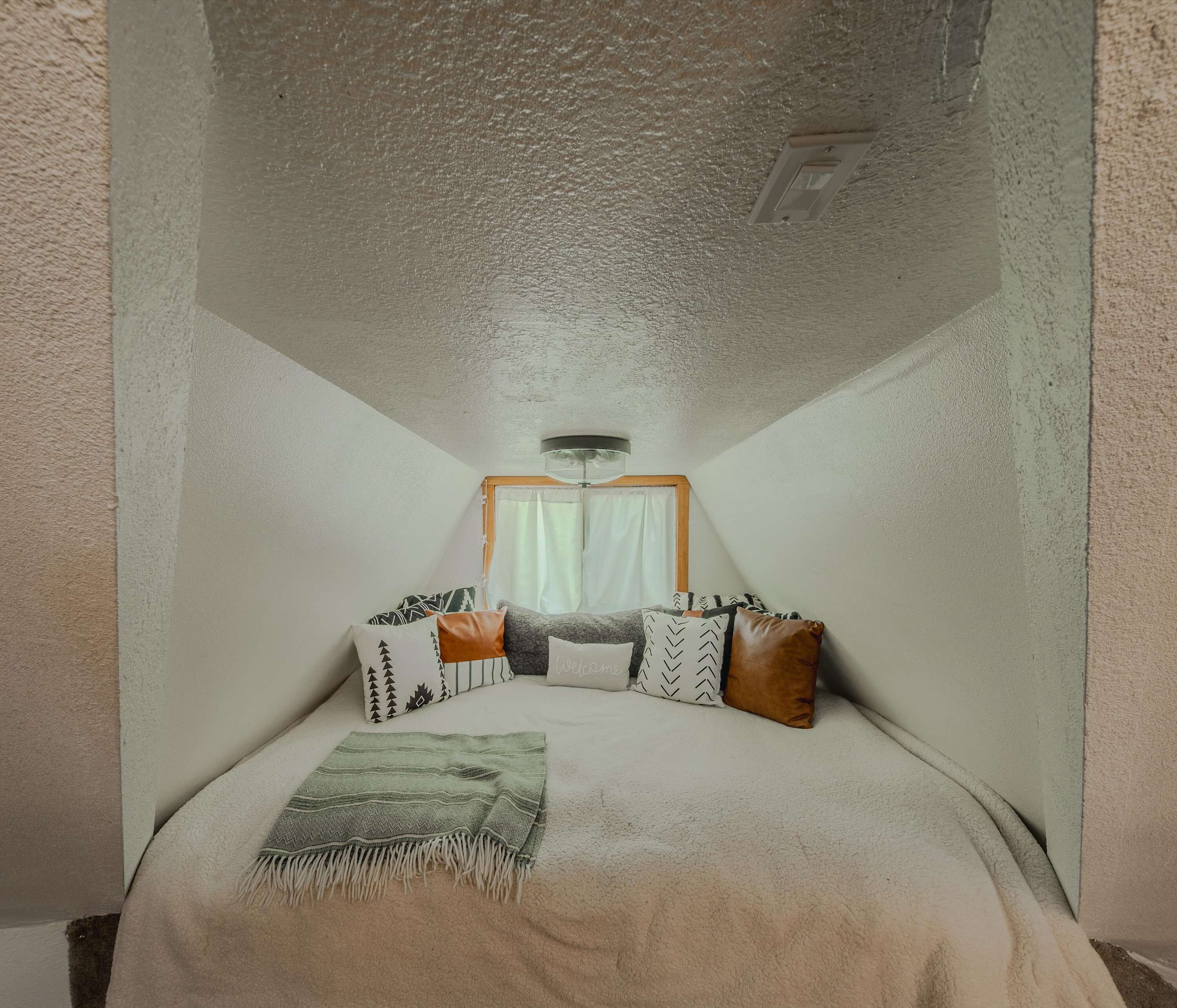 Una stanza piccola e accogliente con un letto, cuscini, una coperta, una finestra con tende e un lampadario.