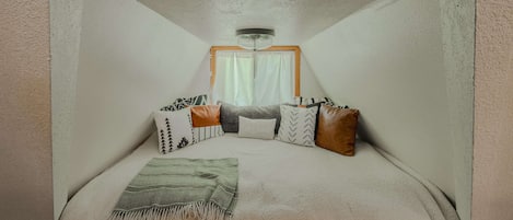 Una stanza piccola e accogliente con un letto, cuscini, una coperta, una finestra con tende e un lampadario.