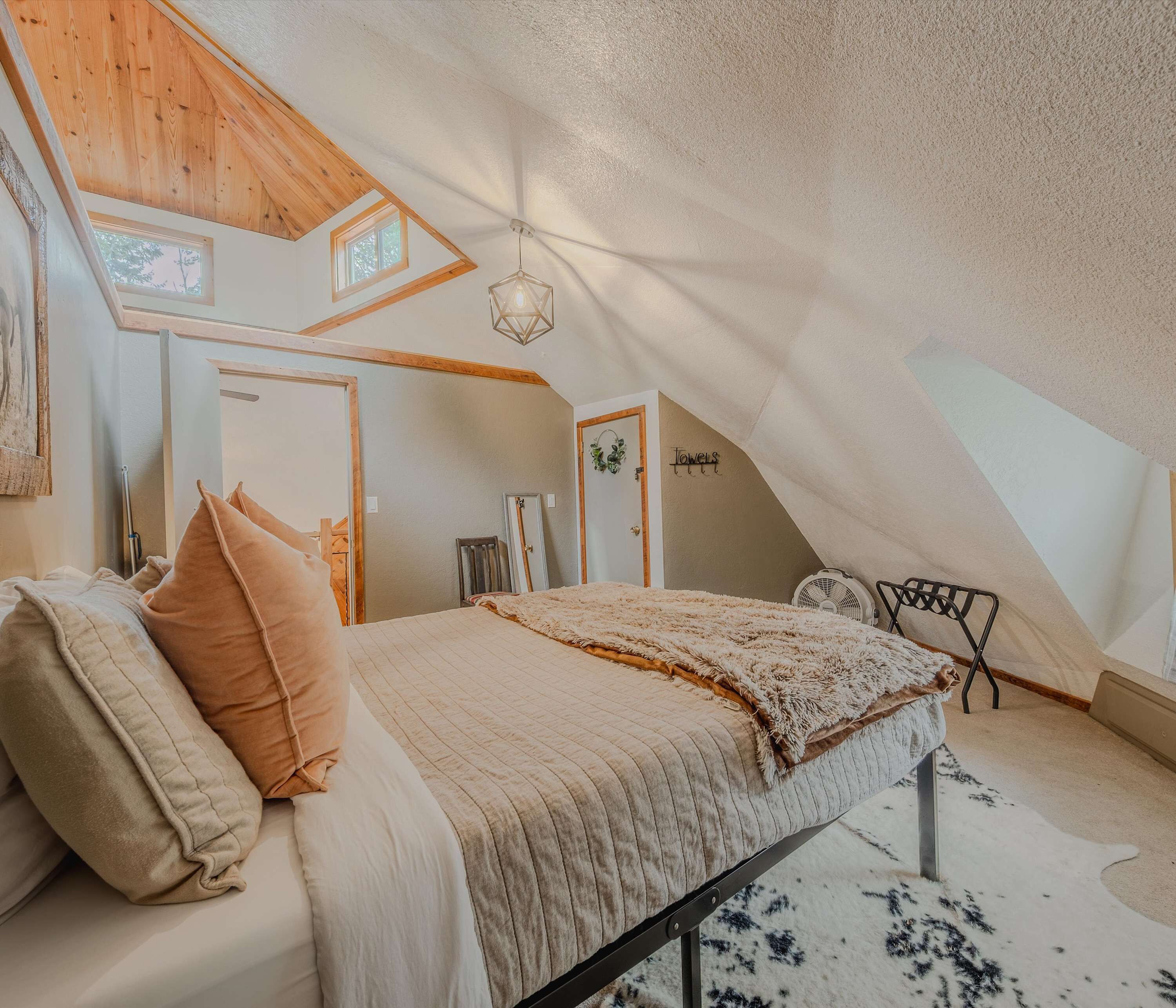 A bedroom with a sloped ceiling, a bed with a fluffy blanket, a hanging light fixture, and a small fan.