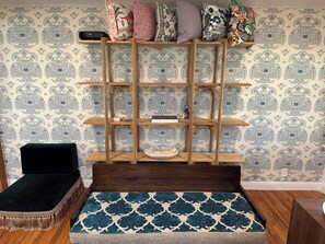 A room with a patterned wallpaper, a wooden shelf unit, a black cushioned bench, and a blue patterned rug.