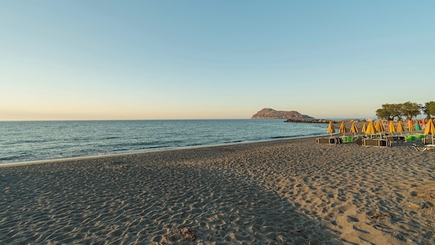A beach with a rocky outcrop in the distance, a row of beach chairs and umbrellas, and a clear sky.