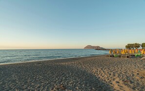 Una playa con un promontorio rocoso a lo lejos, una hilera de sillas y sombrillas, y un cielo despejado.