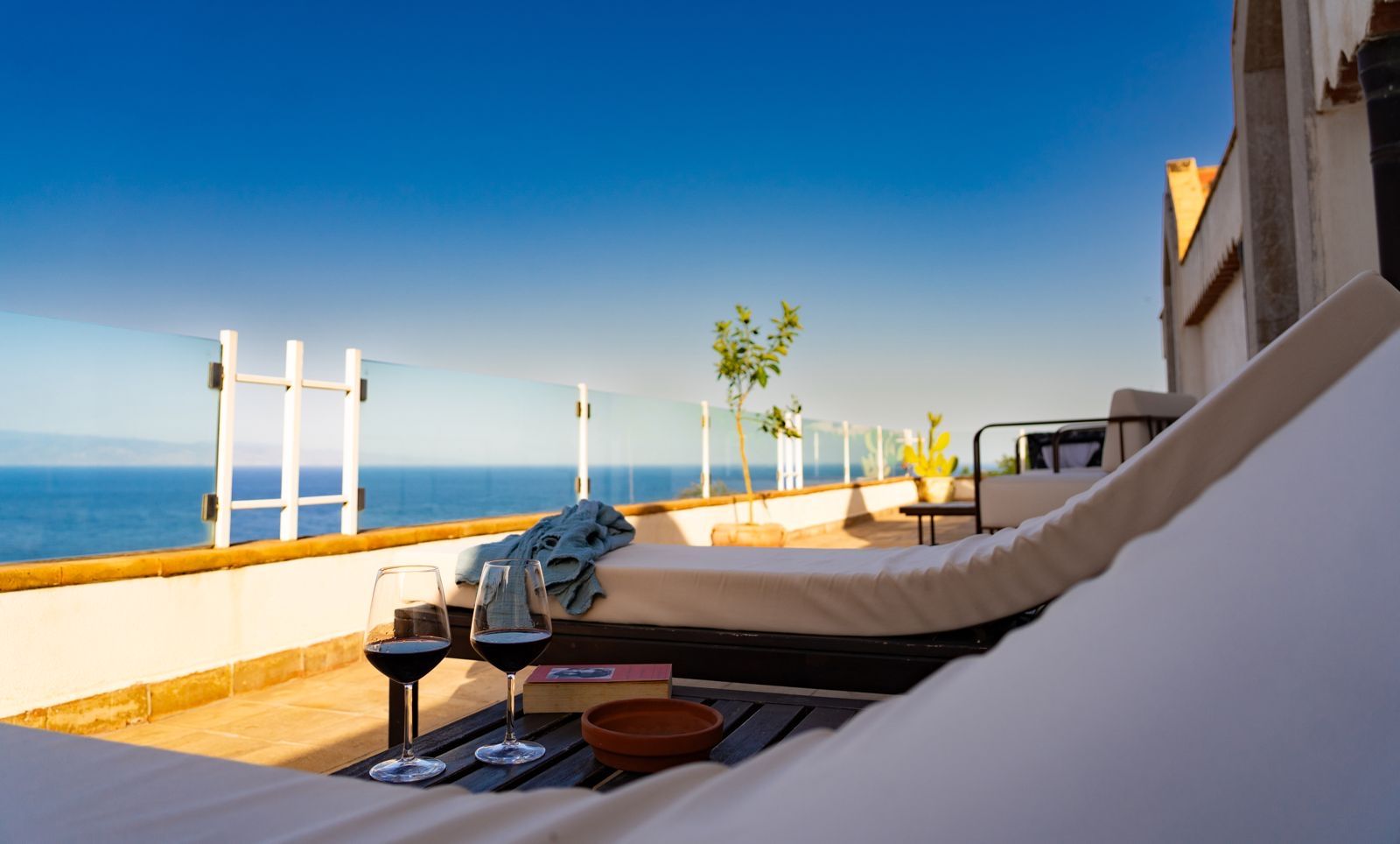 A sunlit balcony with a view of the sea, a lounge chair, a table with a tray, and a potted plant.