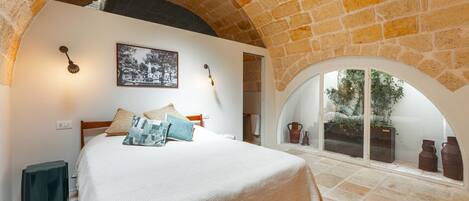 Un dormitorio con techo de piedra, una cama con colcha blanca, un cuadro en la pared y vistas a través de un ventanal de arco.