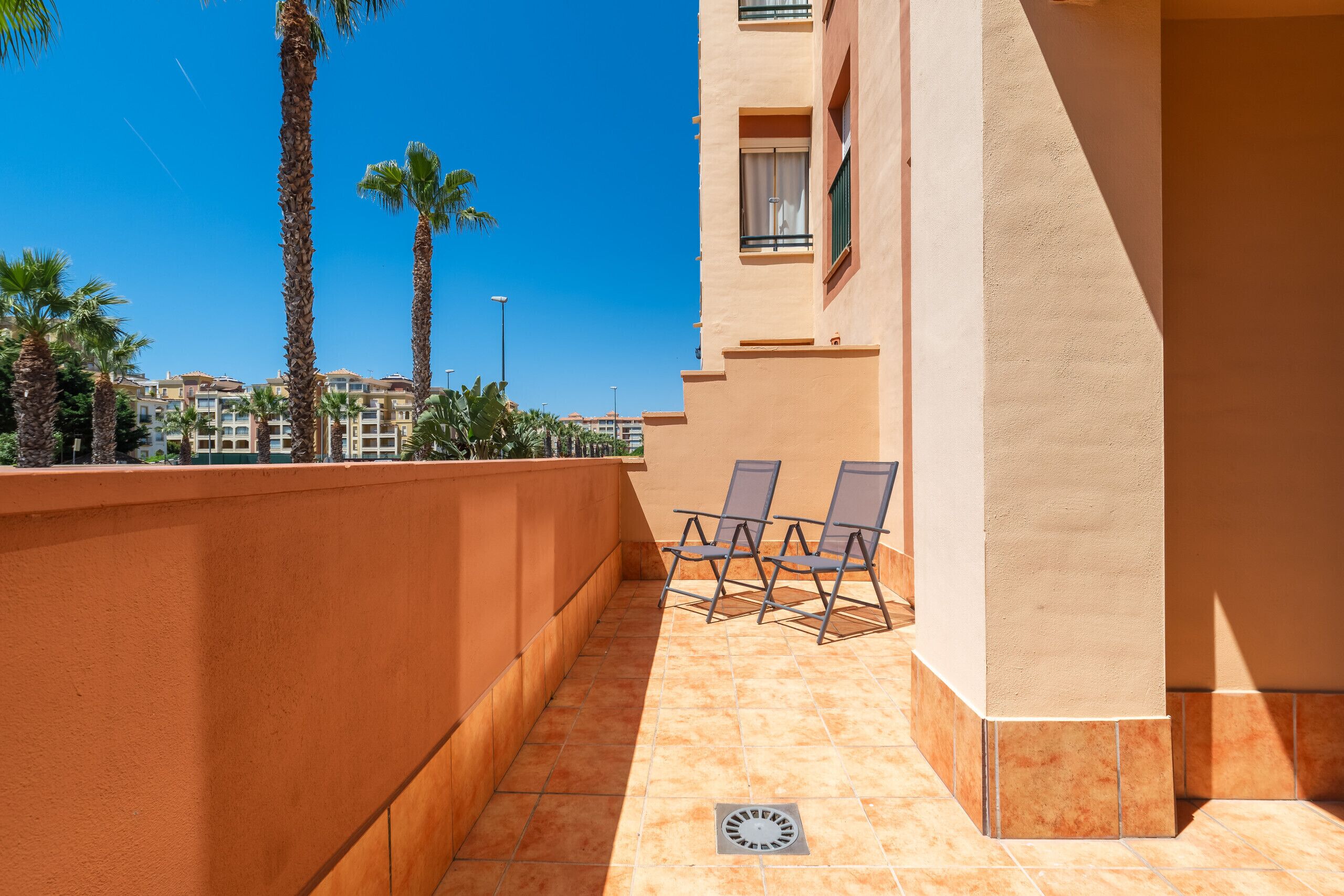 Un balcon avec deux chaises, un sol carrelé et une vue sur des palmiers et des immeubles.