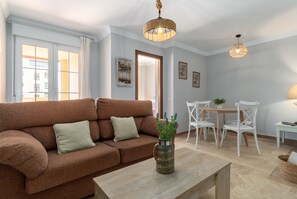 Ein Wohnzimmer mit einem braunen Sofa, einem Holztisch, einem Esstisch mit weißen Stühlen und einer Pendelleuchte.