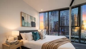 Ein modernes Schlafzimmer mit einem großen Bett, einem Nachttisch, einer Lampe und Blick auf die Stadt durch große Fenster.