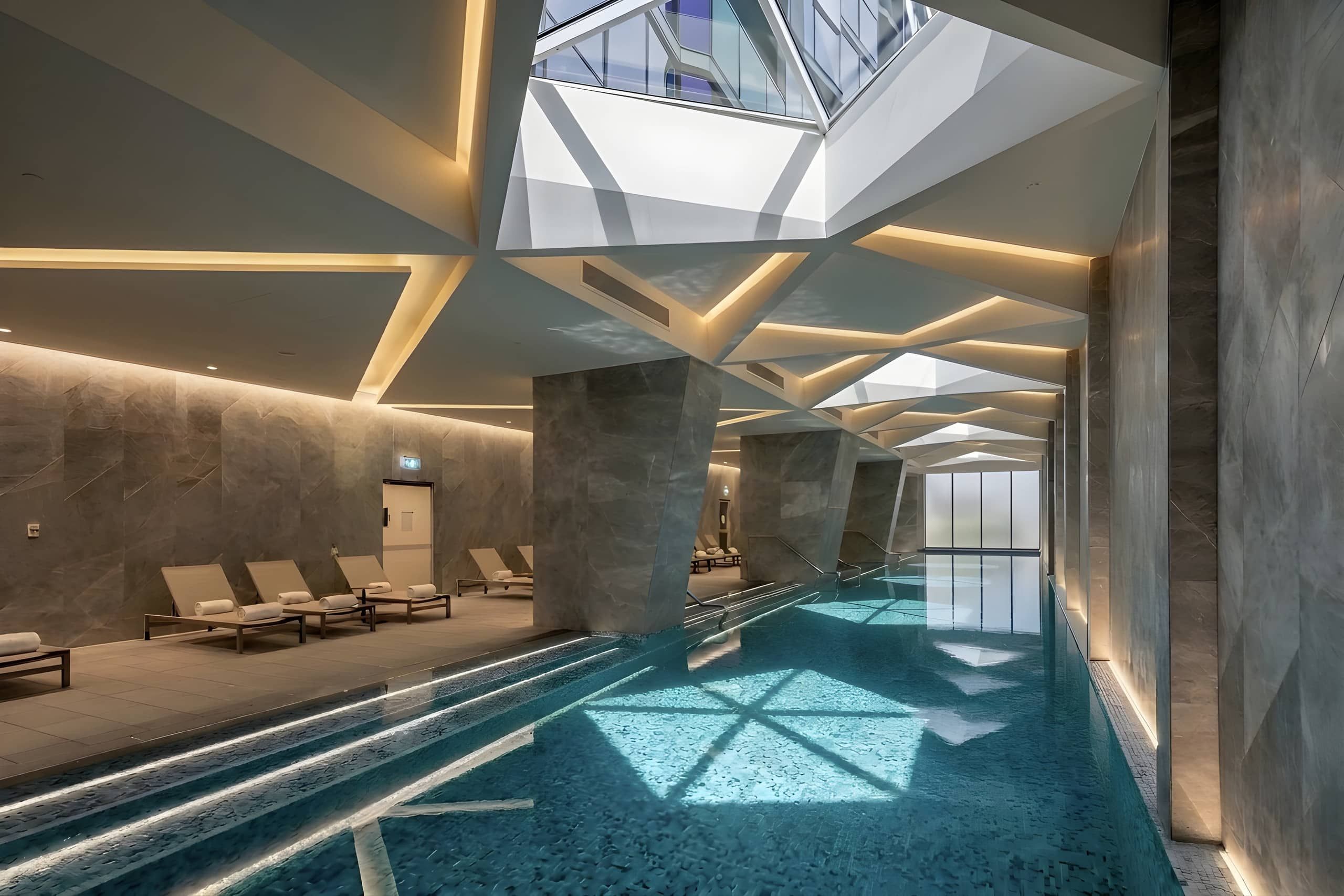 Une piscine intérieure moderne, dotée d’un plafond au design géométrique, d’espaces de détente et d’une verrière.