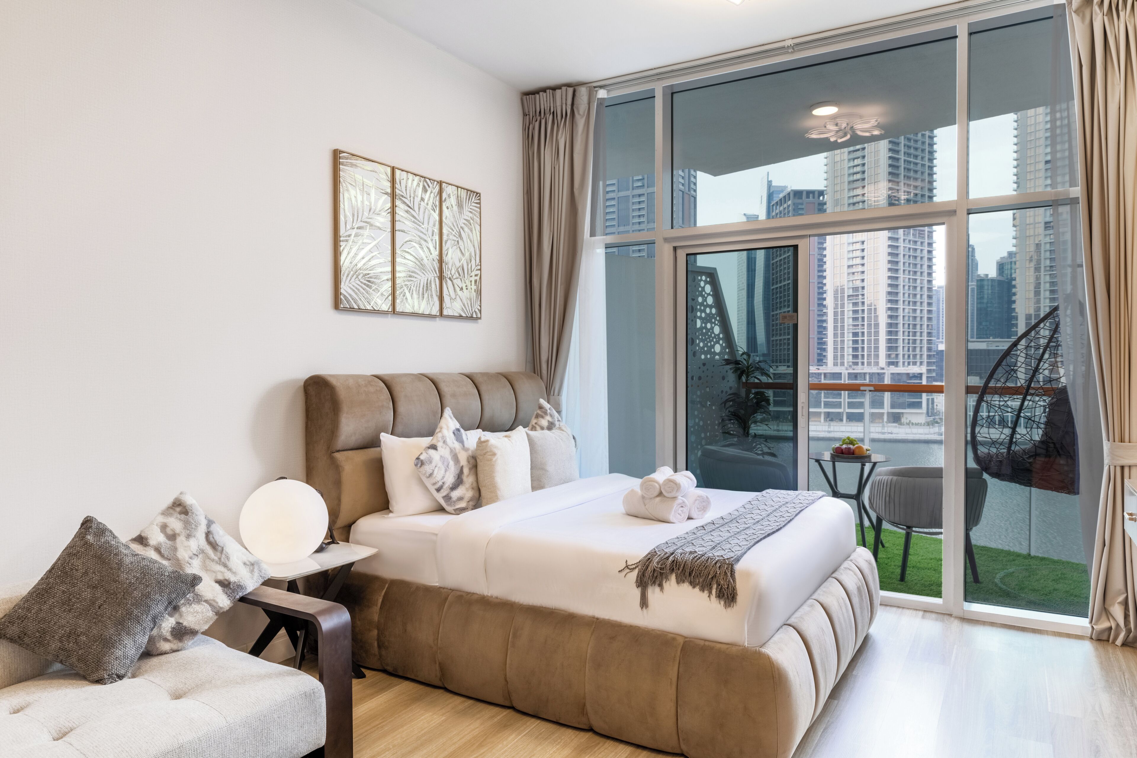 Une chambre moderne avec un grand lit, un canapé, une petite table avec des fruits, et une vue sur le paysage urbain à travers de grandes fenêtres.