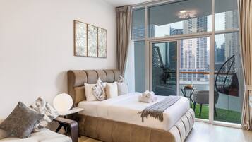 Une chambre moderne avec un grand lit, un canapé, une petite table avec des fruits, et une vue sur le paysage urbain à travers de grandes fenêtres.