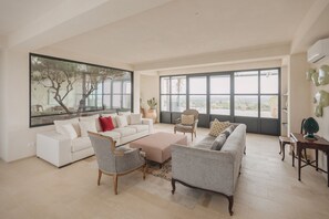 Ein geräumiges Wohnzimmer mit großem Fenster, einem weißen Sofa, einem grauen Sofa, einem Holzstuhl und einem kleinen Tisch.
