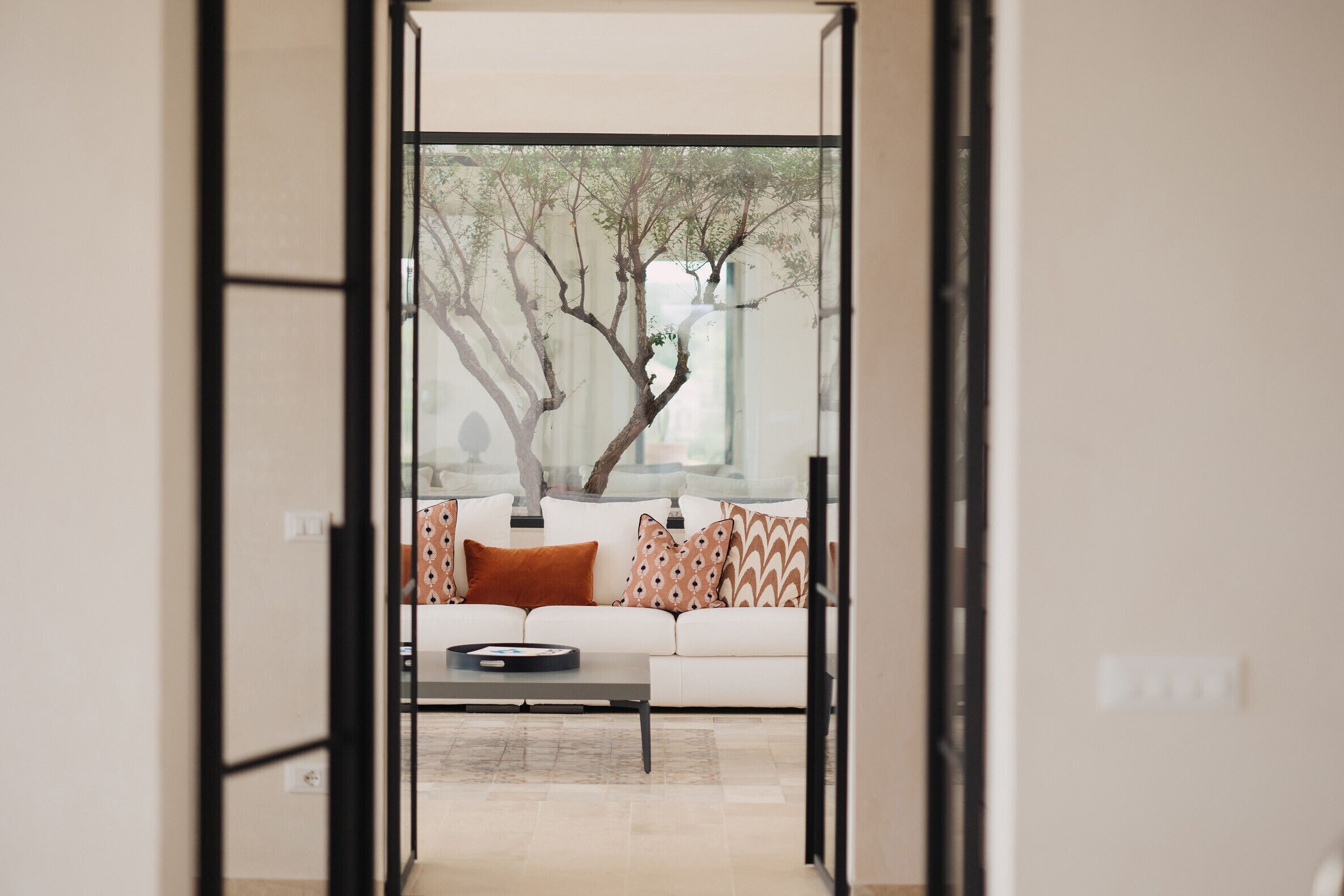 A modern living room with a sofa, patterned cushions, a coffee table, and a view of trees through a glass door.