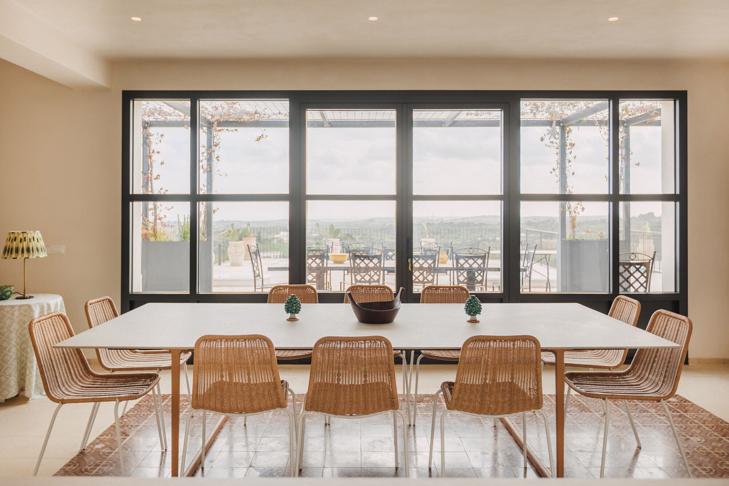 A dining area with a large table, wicker chairs, and a view through large windows.