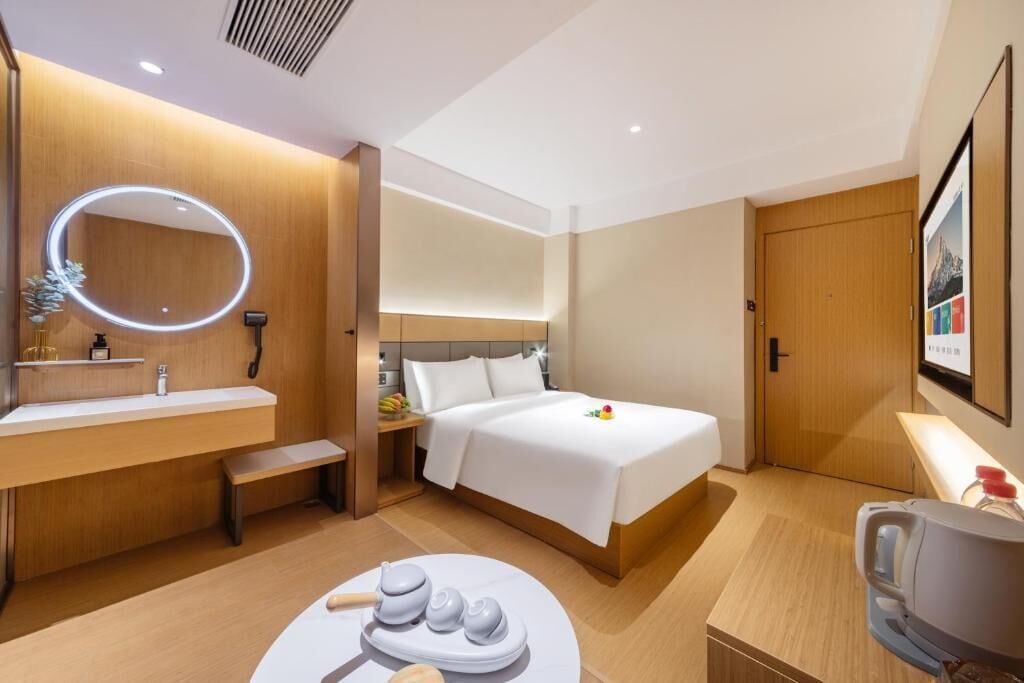A hotel room with a bed, a bedside table, a desk, a chair, a mirror, and a wall-mounted TV.
