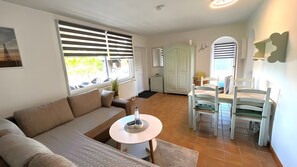 Ein Wohnzimmer mit einer Couch, einem runden Tisch, einem Esstisch mit Stühlen, einem Fenster mit Jalousien und Blick ins Freie.