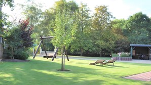 Une zone herbeuse avec une aire de jeux comprenant des balançoires, un espace de repos abrité et un vélo garé près d’un bâtiment.