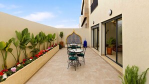 Un patio con mesa y sillas, rodeado de plantas en macetas y un muro decorativo.