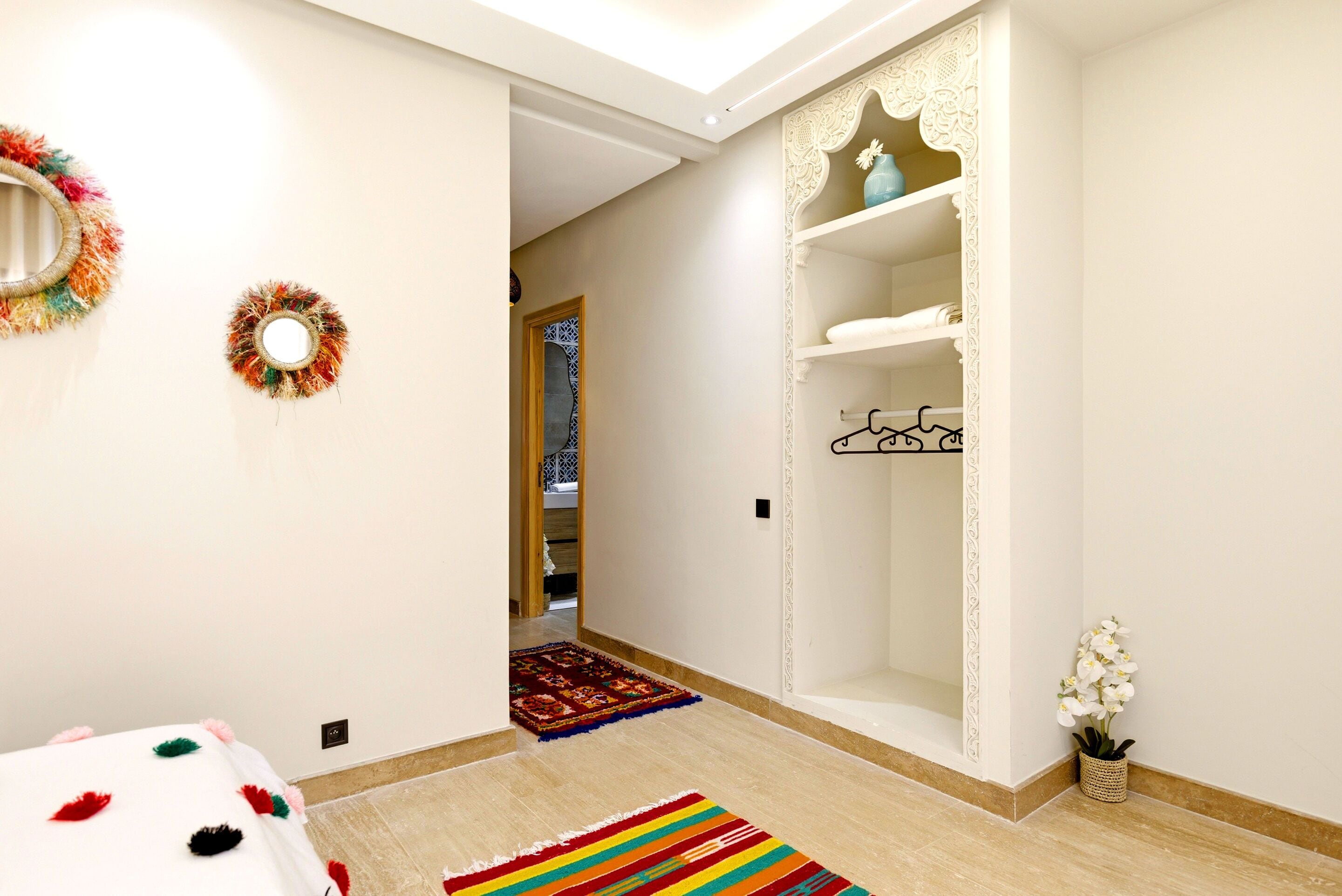 A hallway with a bed, a hanging wreath, a patterned rug, a white shelf unit, and a small potted plant.