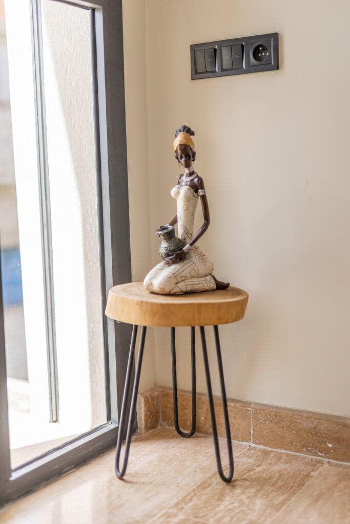 A statue on a wooden stool by a window with a thermostat on the wall.