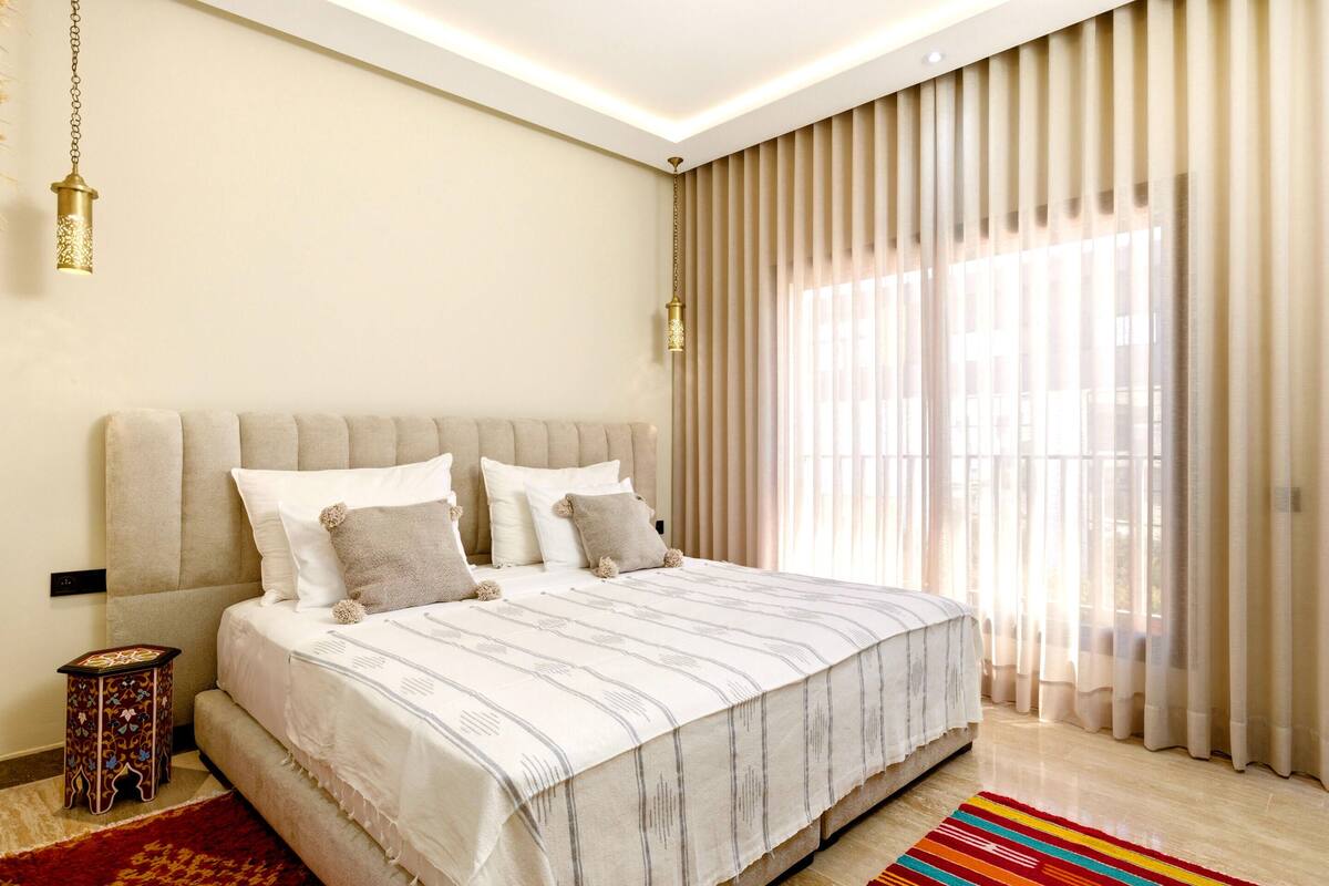 Un dormitorio con una cama grande, una mesita de noche, una lámpara y una ventana con cortinas.