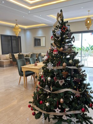 A modern living room with a large Christmas tree, a dining area with a wooden table and chairs, and a large window with a view of greenery outside. - Riad Villa Alain (Deroua)
