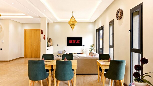 Una habitación de hotel moderna con una mesa de comedor de madera, sillas tapizadas en verde, un ventanal grande y un televisor de pared que muestra 'NETFLIX'.