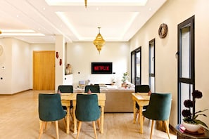 Una habitación de hotel moderna con una mesa de comedor de madera, sillas tapizadas en verde, un ventanal grande y un televisor de pared que muestra 'NETFLIX'.